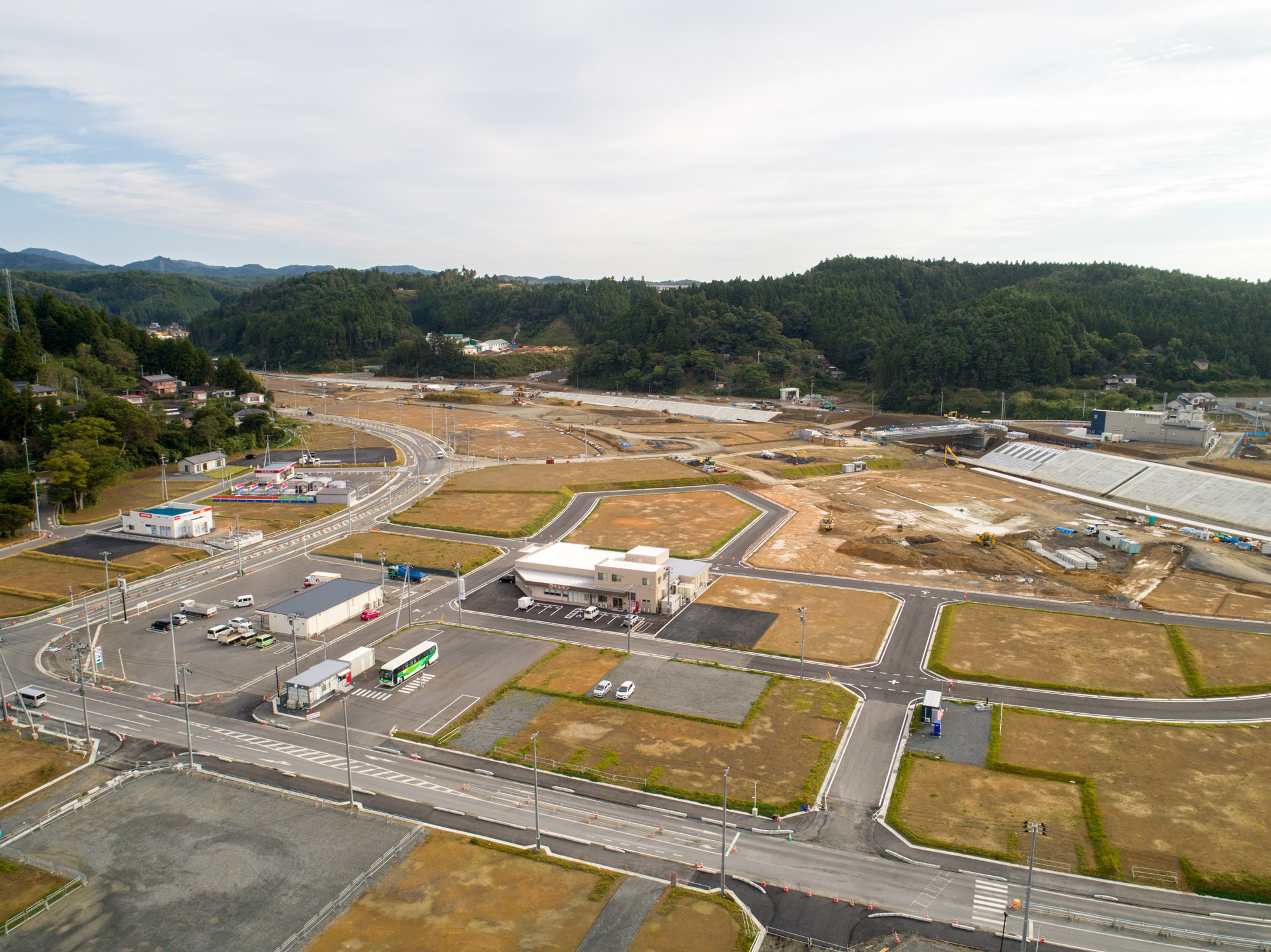 複数の区画や道路、建物が点在し、車両が建物付近に停まっている様子から、開発または建設中であることがうかがえる志津川地区を、緑地や丘を背景に上空から捉えた写真