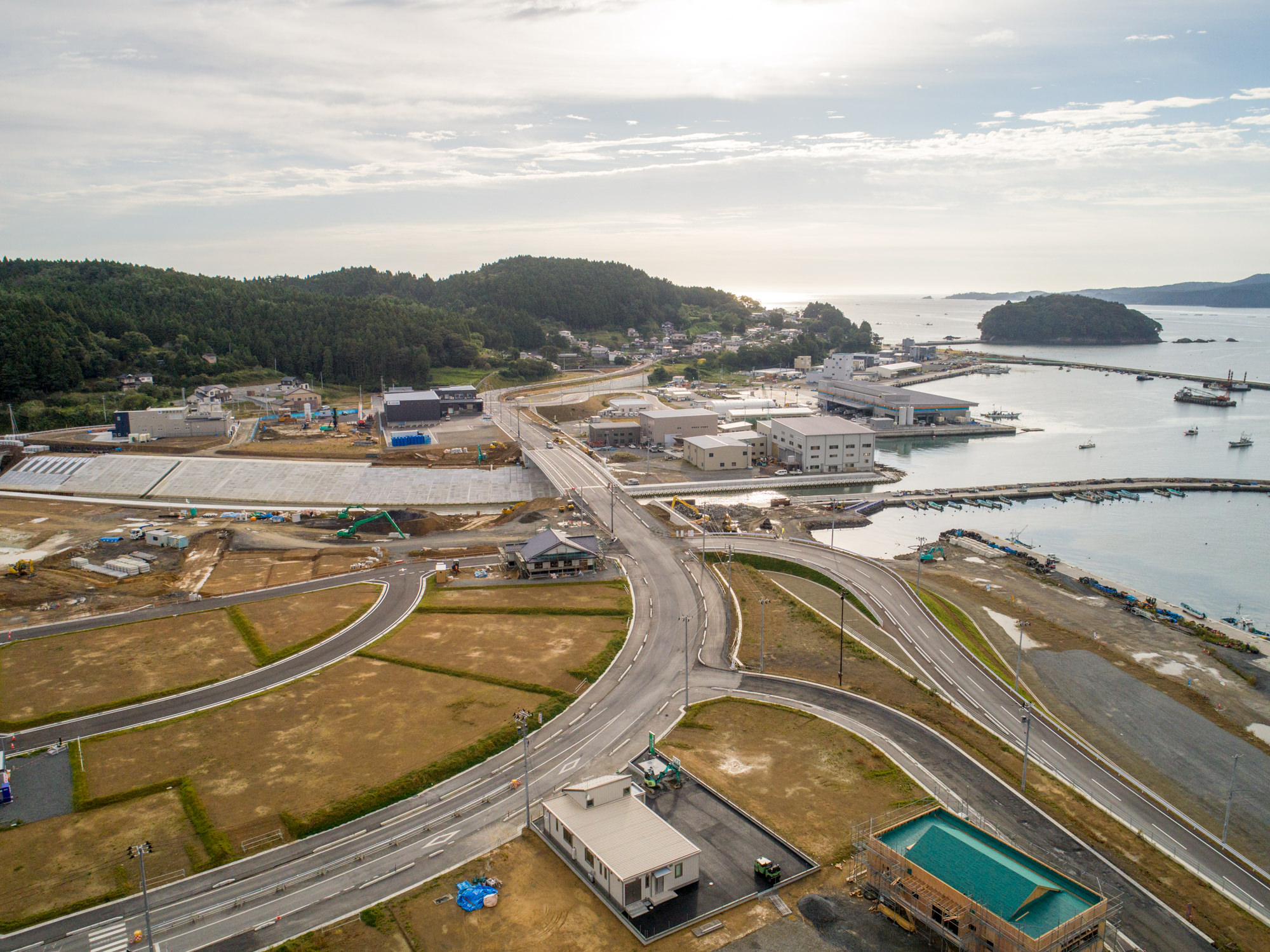 複数の建物や道路が広がり、港には船が停泊している海沿いの町を、木々に覆われた丘に囲まれた風景とともに、太陽の光が水面に反射する晴れ間の空の下で上空から捉えた建設現場の写真
