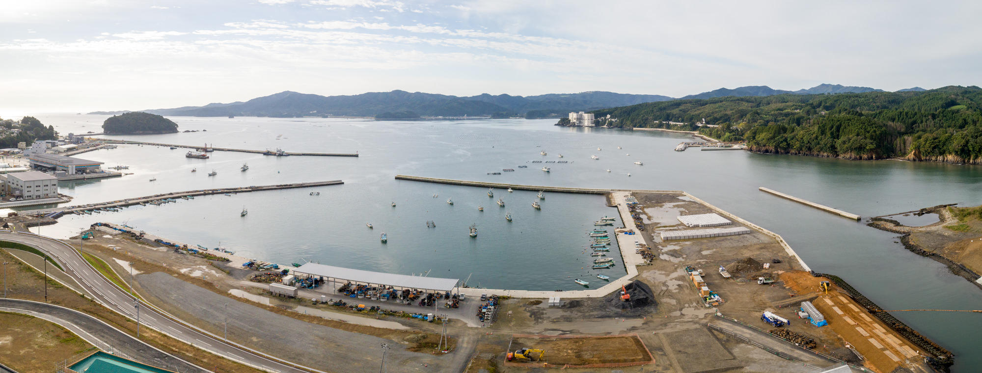 防波堤に囲まれた港に複数の船が停泊し、左側には港湾関連の建物や設備、右側には緑豊かな丘陵地と自然が広がり、水辺近くでは建設作業が行われている様子を、遠くの山々と雲が浮かぶ空を背景に上空から捉えたパノラマ写真