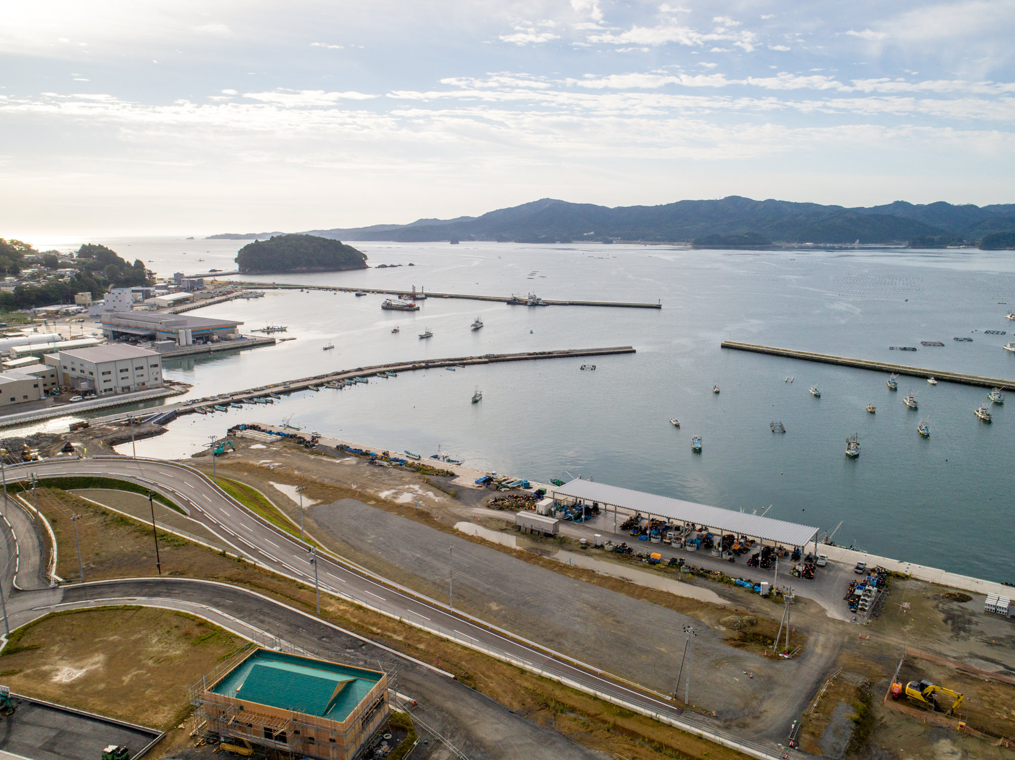複数の桟橋が海に突き出し、多くの船が停泊する港を中心に、建物や道路、広場が広がり、遠くには水面越しに島や丘、山々が見える風景を、雲間から差し込む日差しの下で上空から捉えた建設現場の写真
