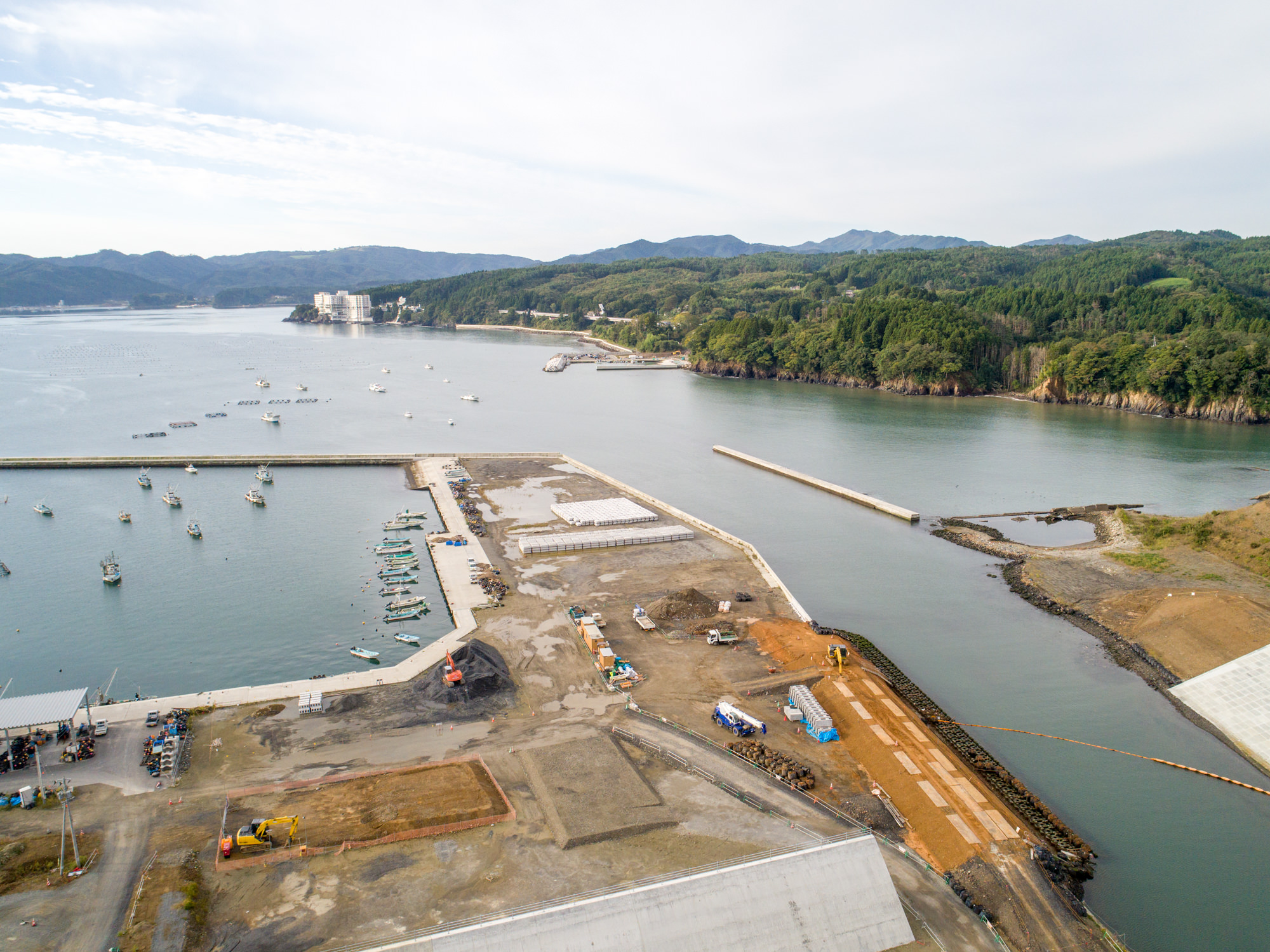 複数の船が桟橋に停泊する港を中心に、水辺に隣接する陸地では建設作業が進行中で、建設車両や機材が見られ、周囲には緑の丘や森林が広がり、海岸沿いには遠くに建物が点在する様子を上空から捉えた写真