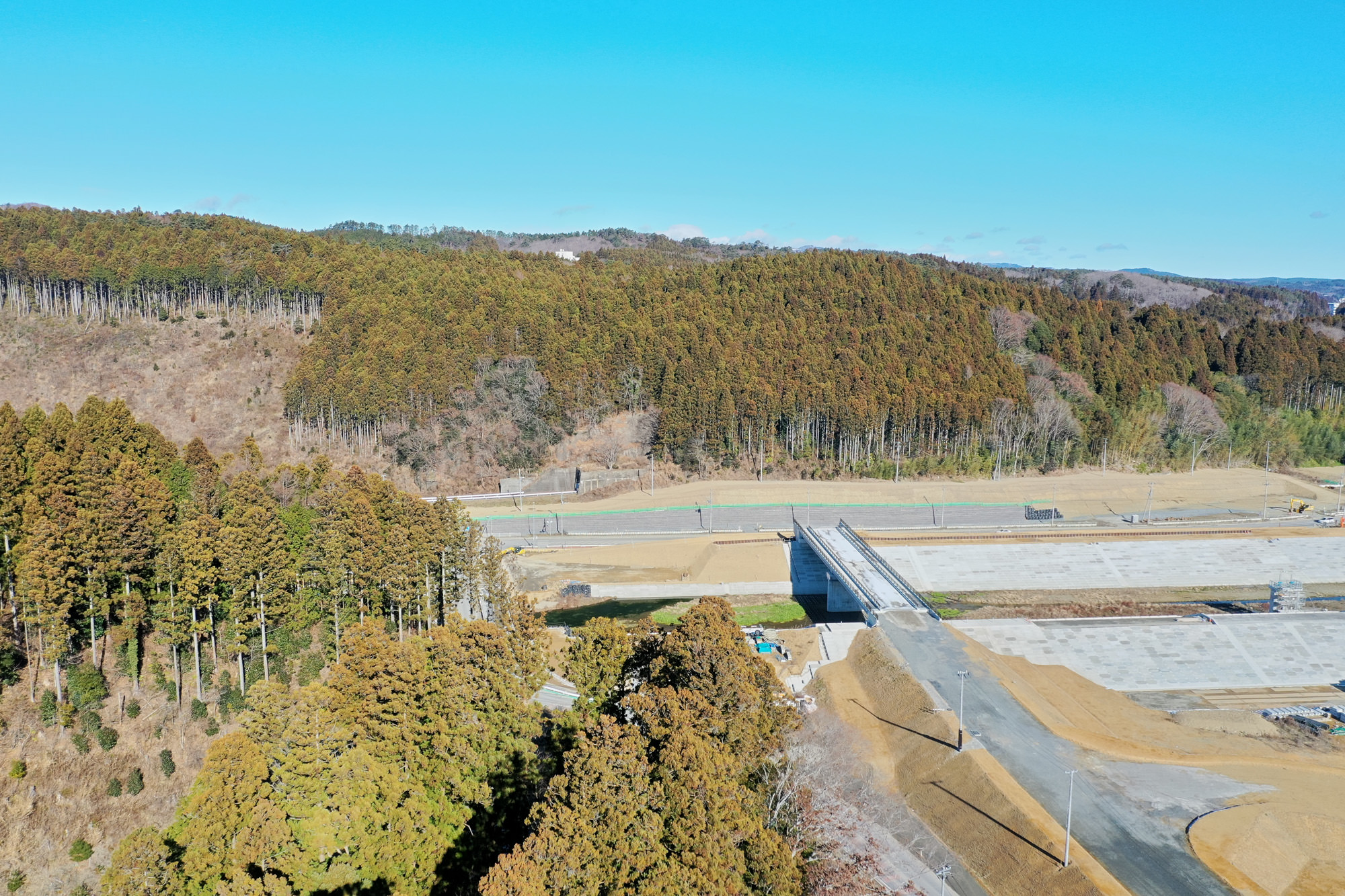 手前に新しく建設されたか建設途中の道路または橋があり、背景には木々が広がる森林が見える静かな風景の写真
