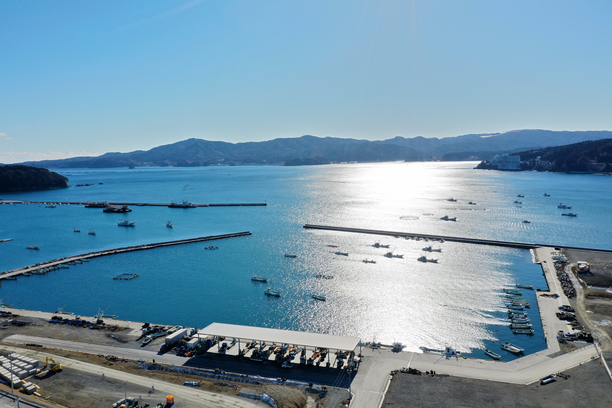 穏やかな水面に太陽の光が反射する港には複数の船が浮かび、桟橋が水中に延び、岸辺には建物や車両が並び、背景には山々が広がっている海辺の風景の写真