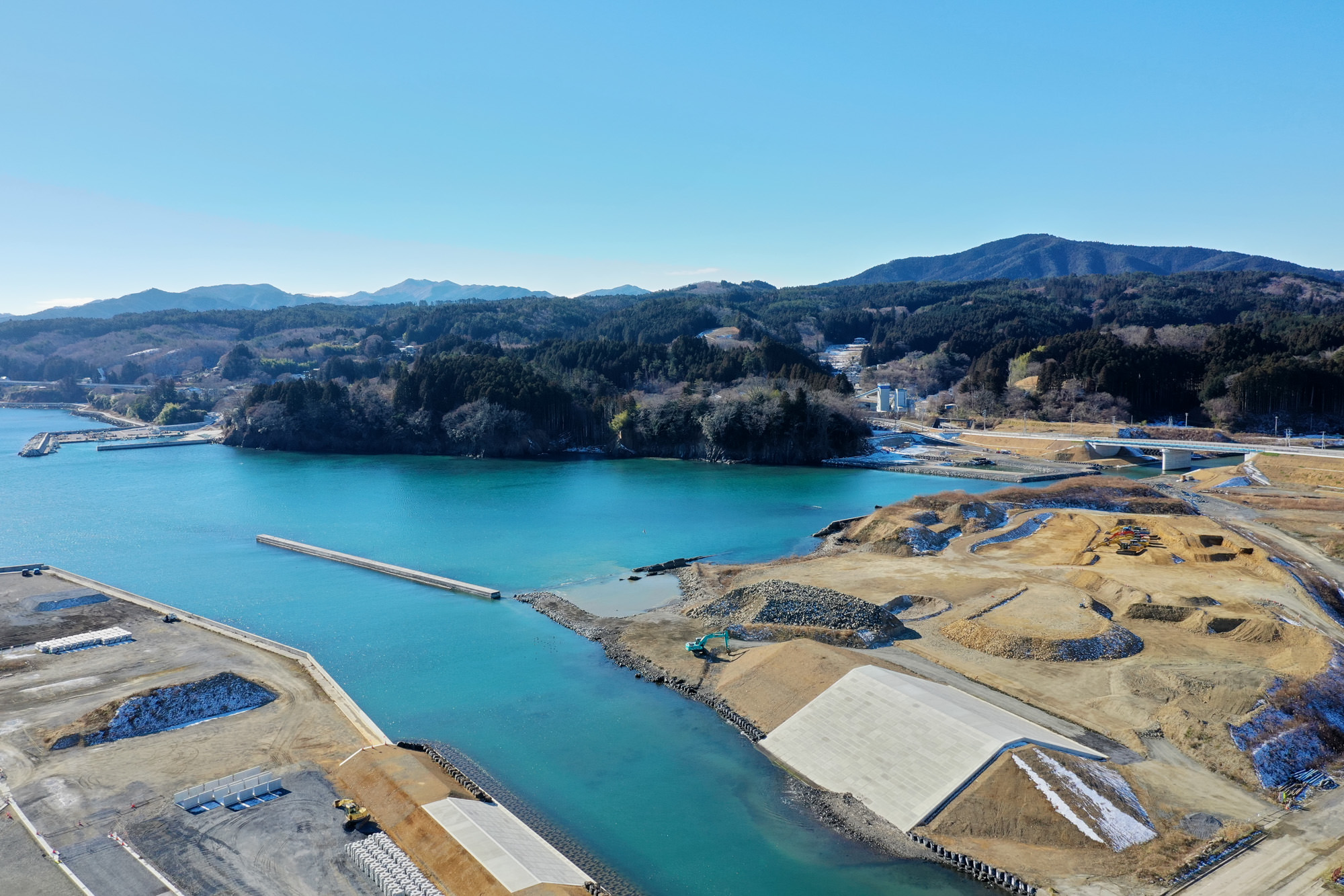 澄んだ青い海に囲まれた海岸地域で、陸地の一部では開発や工事が進められており、背景には木々に覆われた丘や山が広がり、空は晴れ渡っている風景の写真