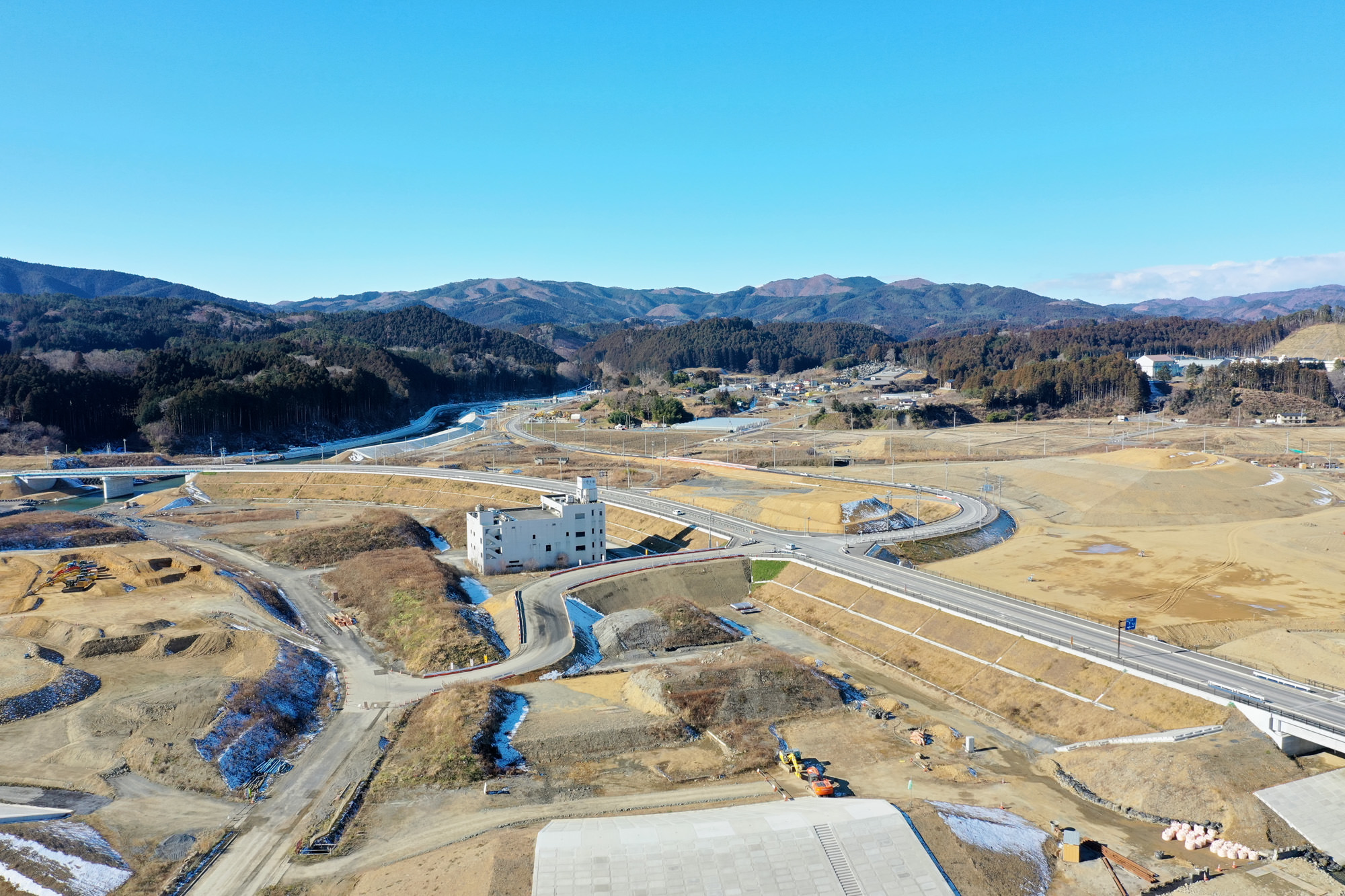 中央に大きな建物があり、その周囲に道路や通路が広がる開発中または工事中の地域で、地面には雪が点在し、背景には山々がそびえ、晴れ渡った青空が広がっている風景の写真