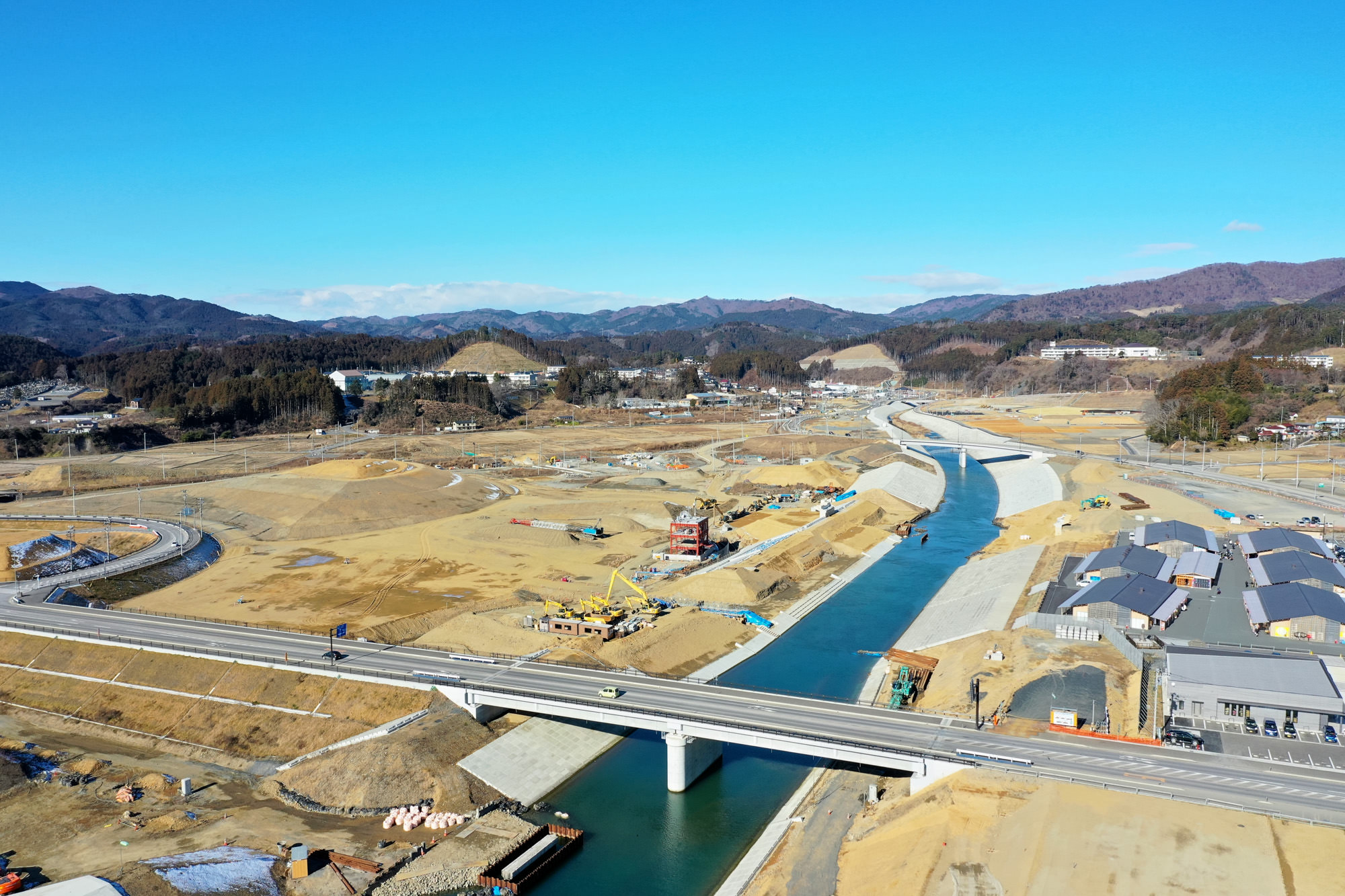 川が中央を流れる工事現場を上空から見下ろし、橋が川を横断し、その周囲には建設機械や資材が点在し、背景には丘や山々が広がり、雲ひとつない青空が広がっている風景の写真