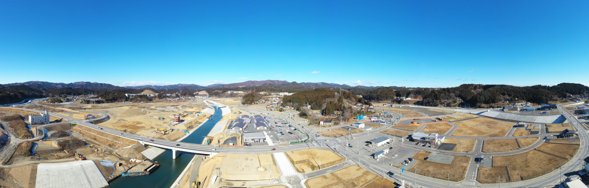 中央に川が流れ、その両側に建物や道路が広がる工事現場を広く見渡すことができ、周囲には木々と丘が広がり、青空が広がっている風景の写真