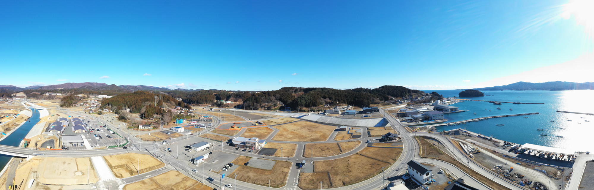海沿いの町を上空から見下ろし、建物や道路が広がる市街地と空き地が混在し、背景には丘が連なり、青空が広がっている風景の写真