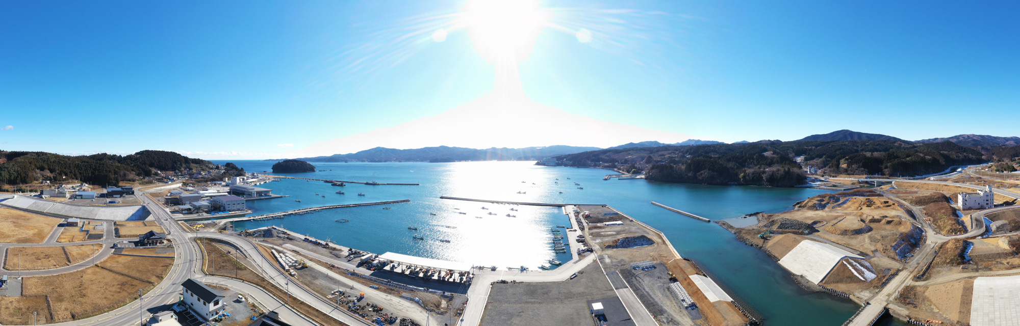 晴れ渡った青空の下、穏やかな海に複数の船が停泊する港が広がり、海岸沿いには建物や構造物が並び、道路が各所へと延び、背景には丘や山々が連なる風景の写真