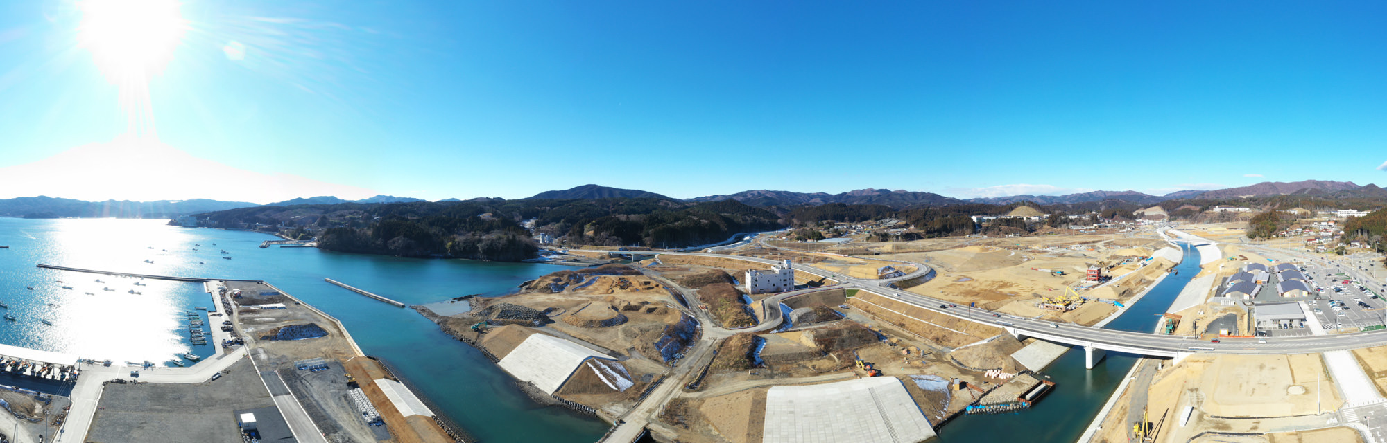 左側に海が広がり、複数の桟橋と船が見える海岸の工事現場では、道路や橋などのインフラ整備が進められ、土木作業が行われており、背景には丘や山々が連なり、晴れ渡った青空の下で太陽が輝いている風景の写真