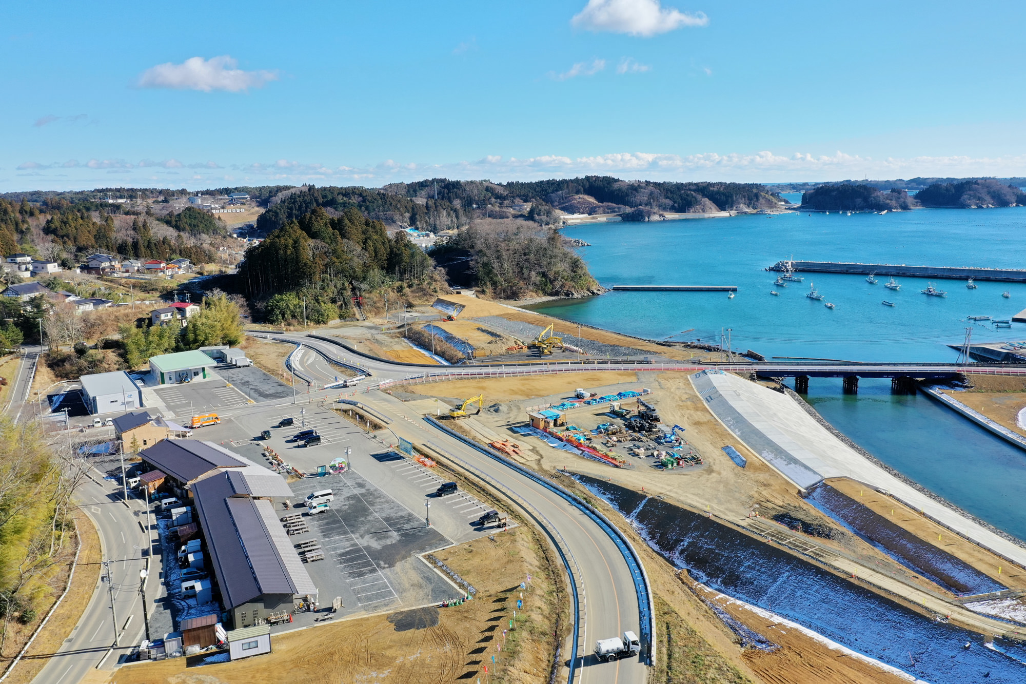 海に面した地域を上空から見下ろし、湾には船が停泊し、入り江には橋が架かり、海岸近くの工事現場には複数の建物や道路、車両があり、背景には森林と丘が広がっている風景の写真