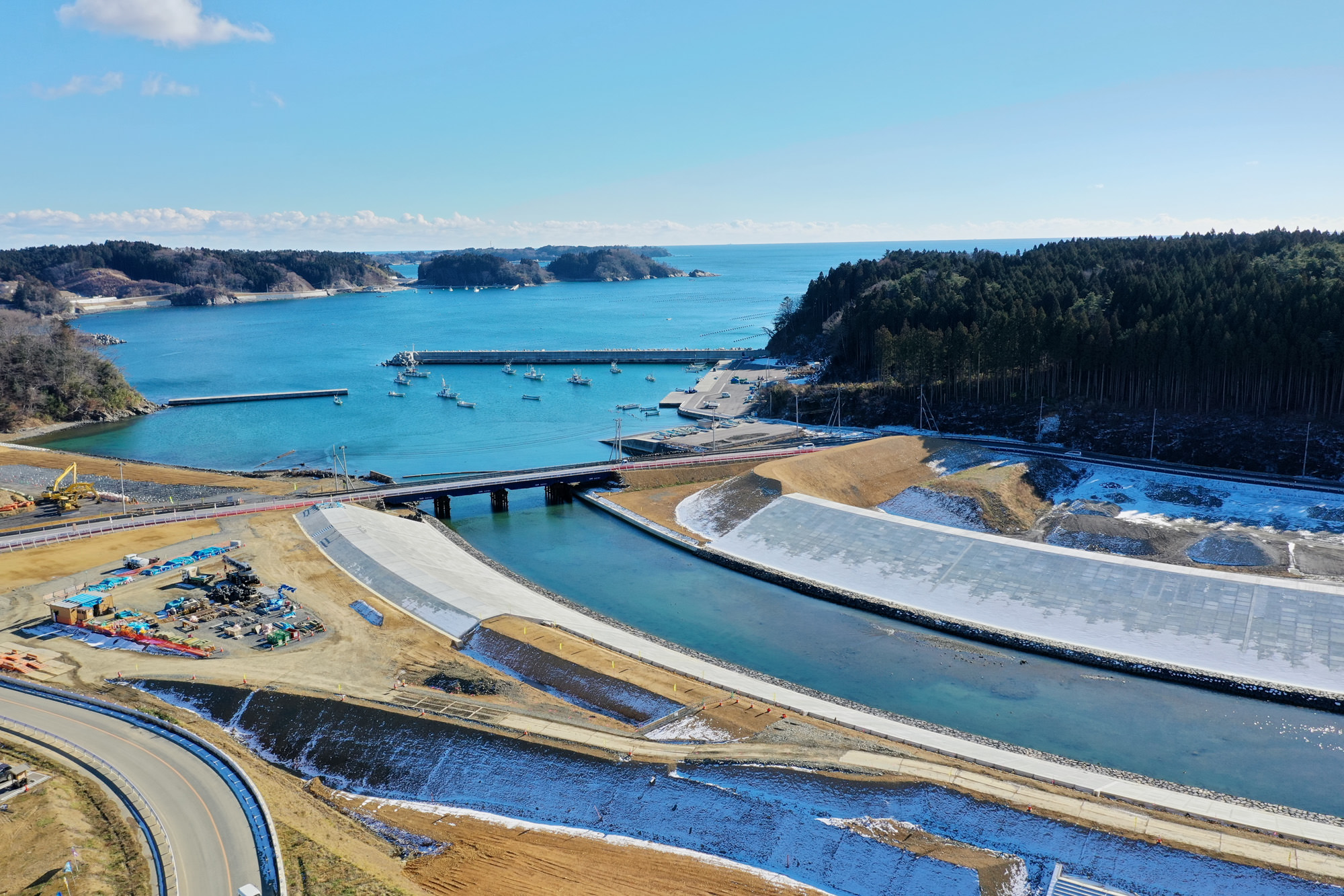 湾または港と思われる水域に橋が架かり、奥には複数の船が停泊しており、左側には建設機材や資材が見られ、右側には森林が広がり、晴れ渡った空が広がっている海岸地域の風景の写真