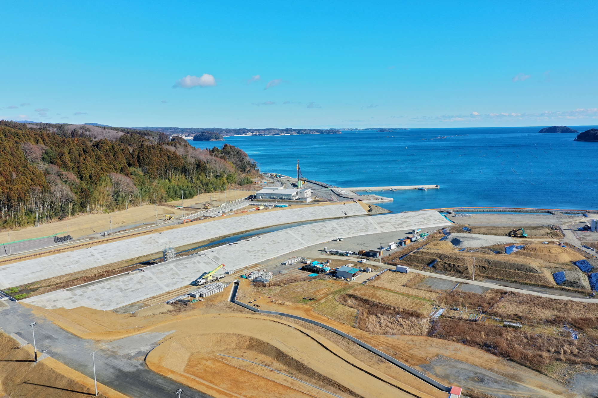 海に面した工事現場で、大きなコンクリート構造物が海岸沿いに建設されており、周囲には木々に覆われた丘と海が広がり、複数の建設車両や仮設施設、道路が点在し、空には少し雲が浮かぶ風景の写真