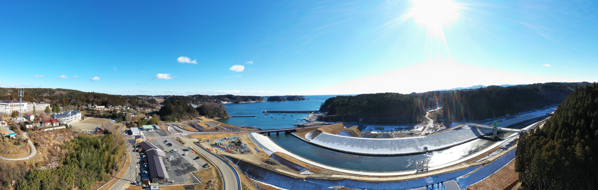 中央に川または運河が流れ、複数の橋がそれを横断し、周囲には森林や丘、建物、道路、駐車場などのインフラが広がり、背景には広大な海が見え、右上には太陽が輝き、空には少し雲が浮かぶ海岸地域の風景の写真