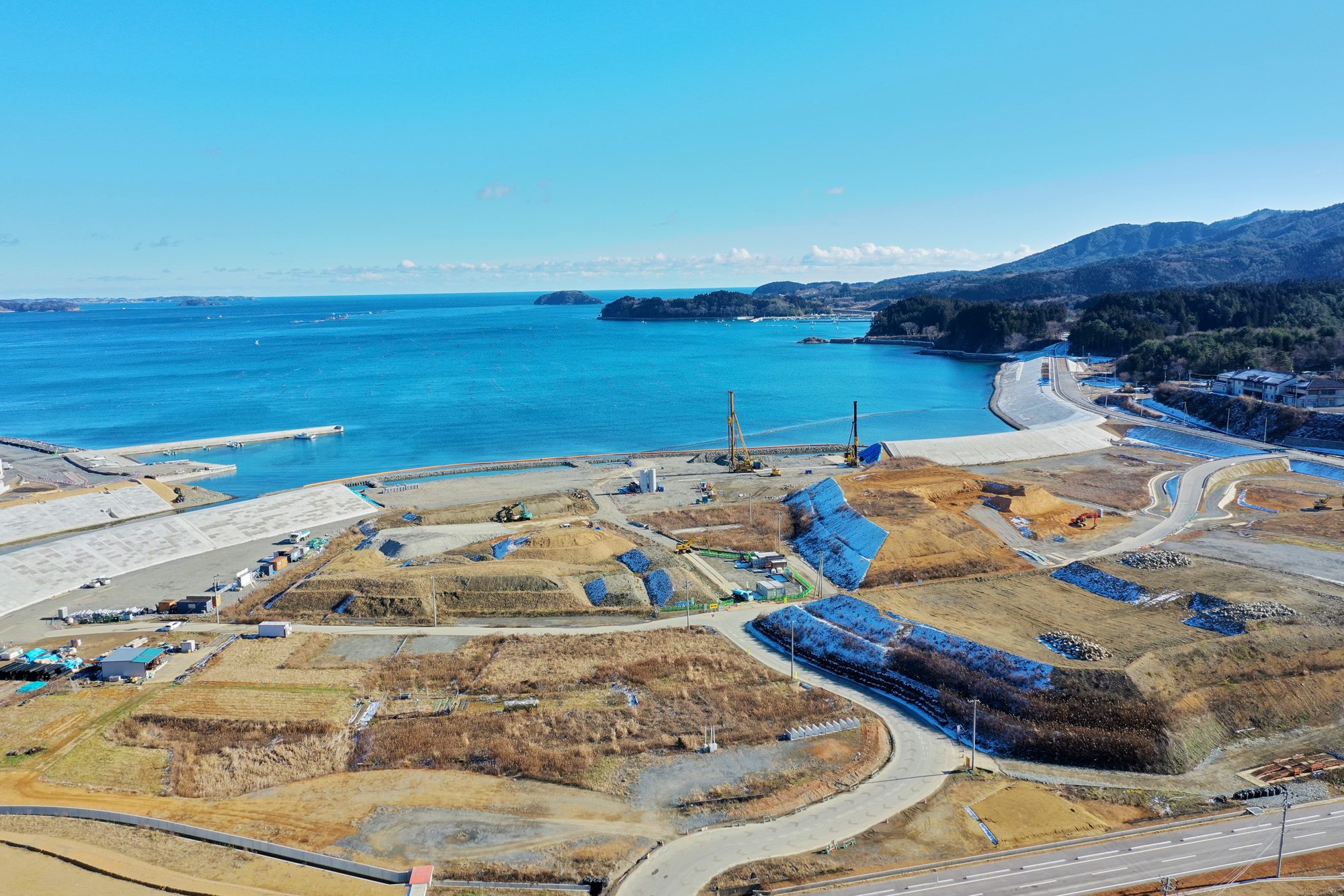 海に面した工事現場で、土砂の山や建設機械、建設途中の構造物が点在し、背景には木々に覆われた丘や山が広がり、上空には青空が広がっている風景の写真