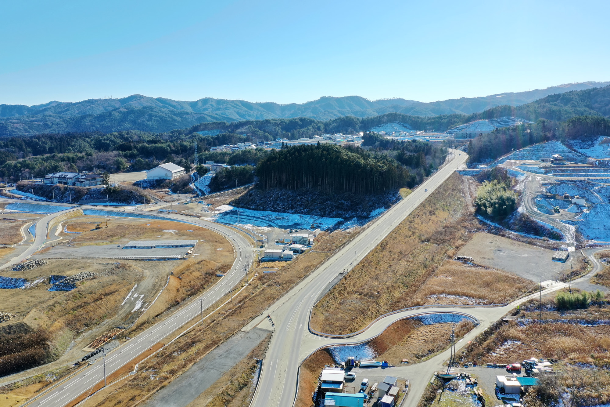 曲がりくねった道路と点在する建物が広がる農村風景を上空から捉え、所々に雪が残り、背景には山々がそびえ、青空が広がっている風景の写真
