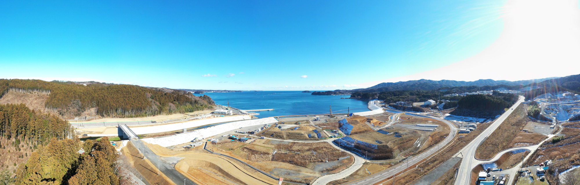 青い空の下、湾に沿って曲がる海岸線に森林、工事現場、道路が広がり、工事区域にはさまざまな構造物や車両が点在し、背景には丘や山々が連なる広大な風景の写真