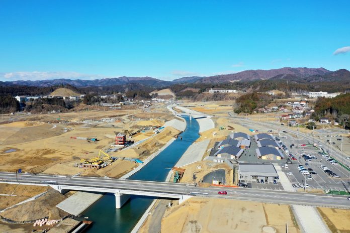 中央に川が流れ、その上に橋が架かり、広範囲にわたる建設現場の中に建物や車が点在している様子が写された風景の写真