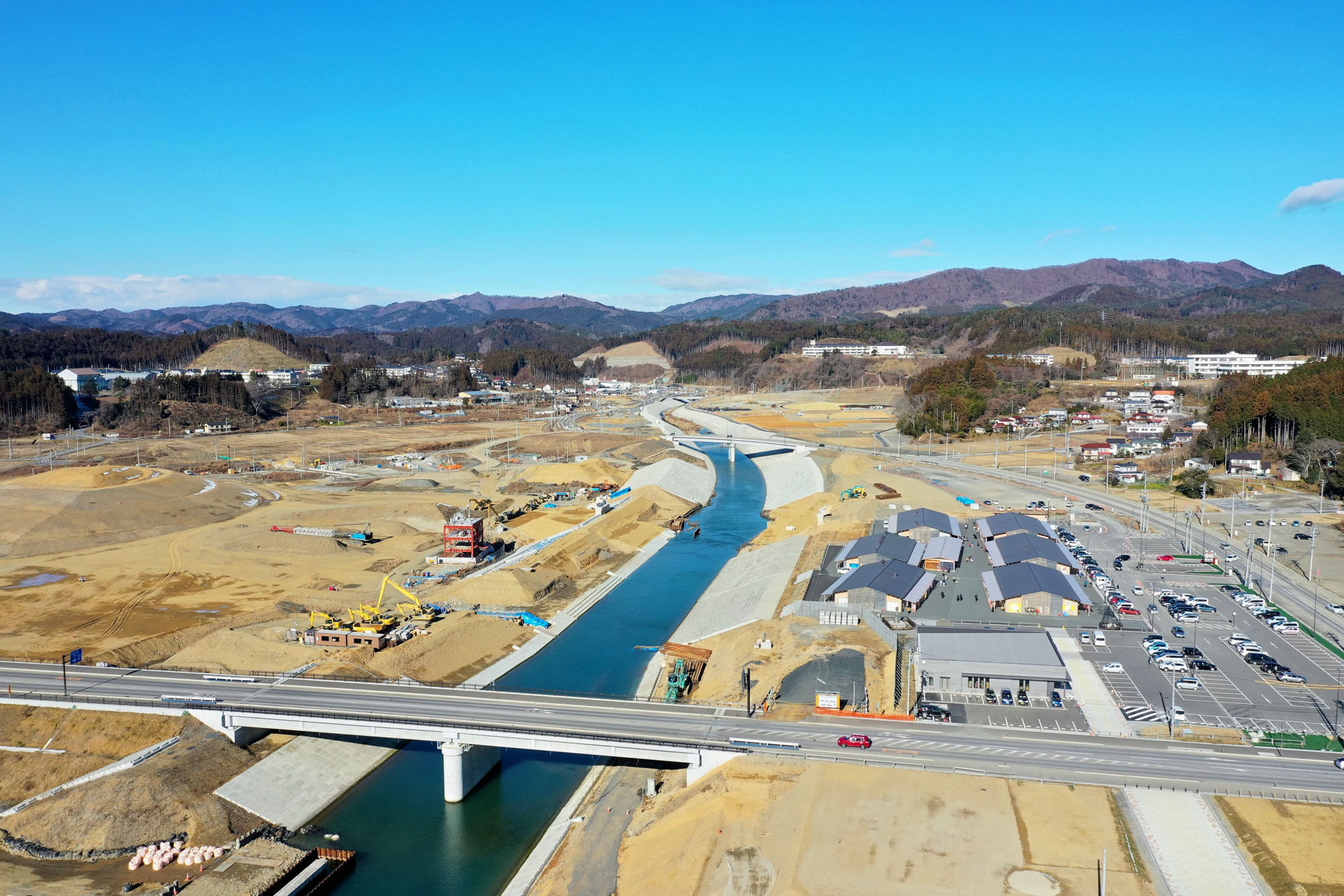 中央に川が流れ、手前には橋が架かり、右側には複数の建物や車両が並び、背景には山々と青空が広がっている工事現場の風景の写真