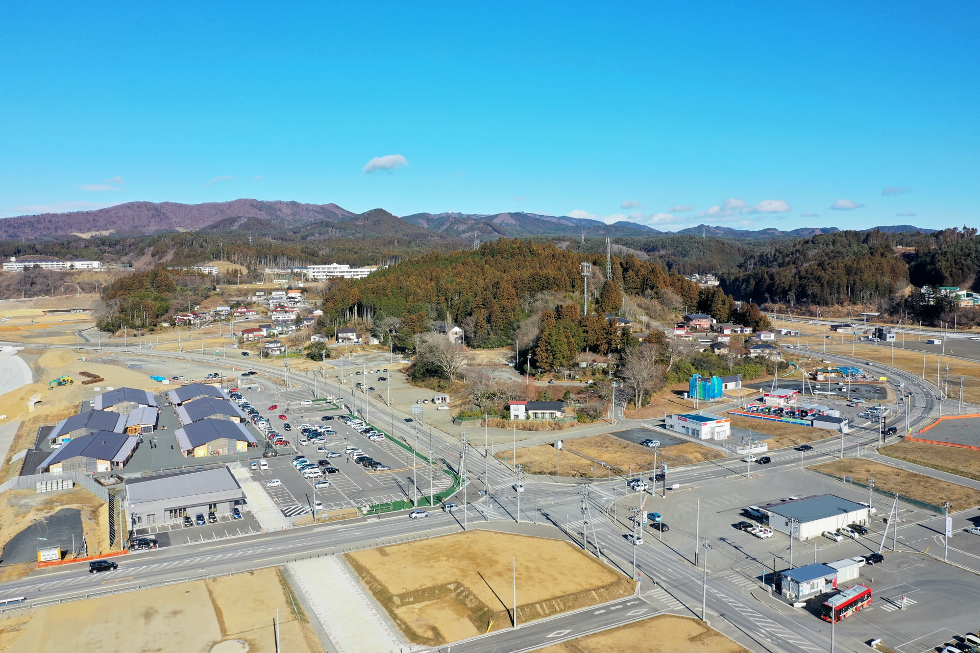 建物や道路、駐車場が広がる場所を上空から見下ろし、複数の車が駐車や走行しており、周囲には草地や森林、背景には丘が広がり、空には少し雲が浮かぶ風景の写真