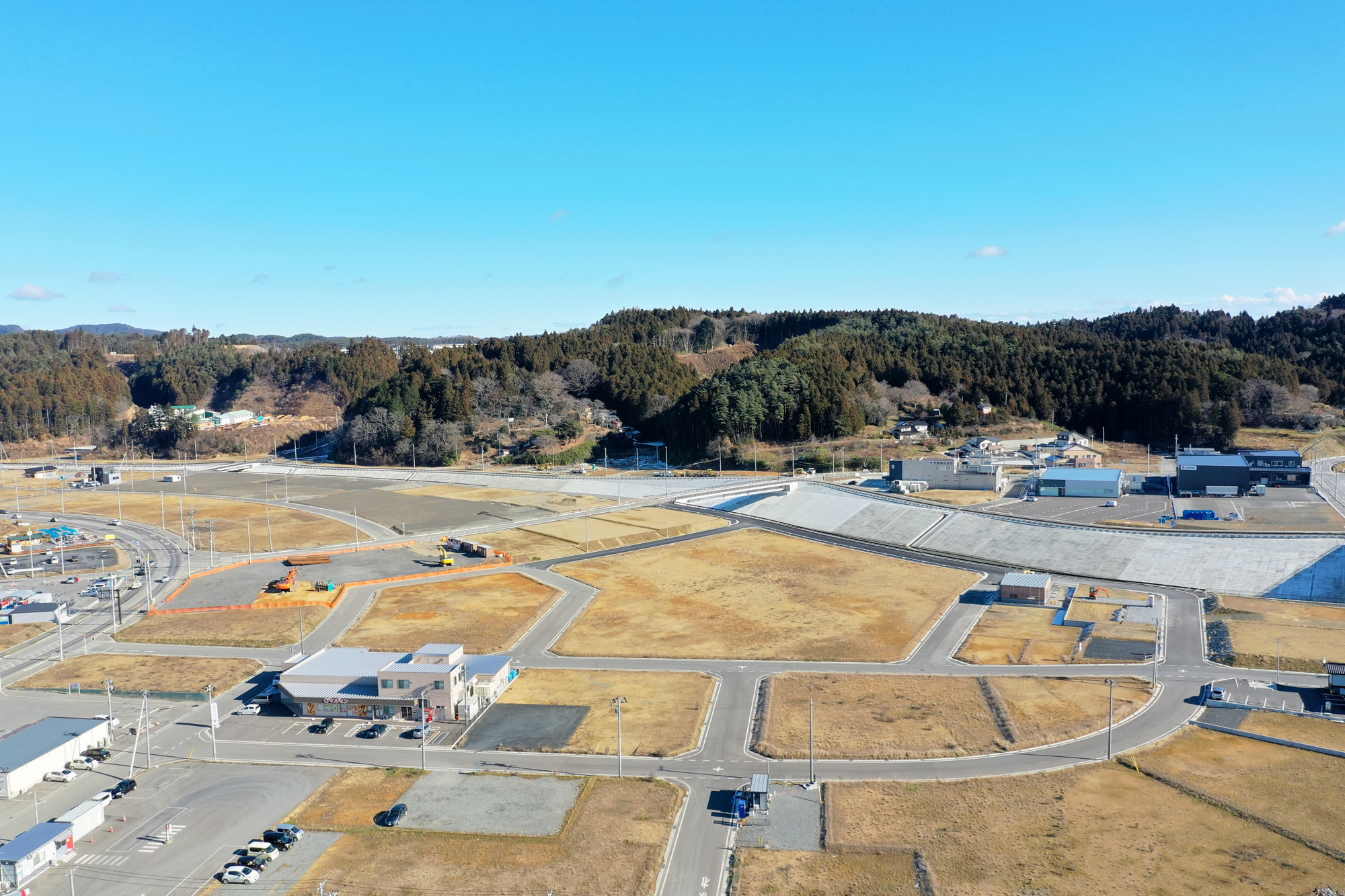 道路で区切られた複数の区画が広がる半農村地域を上空から見下ろし、所々に建物や構造物が点在し、道路には車両が見られ、背景には森林に覆われた丘と青空が広がっている風景の写真