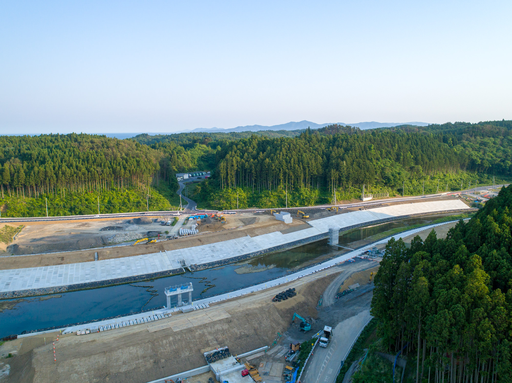 森林に囲まれた地域で進行中の橋または道路の建設現場を上空から撮影したもので、水域をまたぐ構造物があり、建設車両や機材が見られ、周囲には木々が密集し、背景にはなだらかな丘と晴れた空が広がる風景の写真