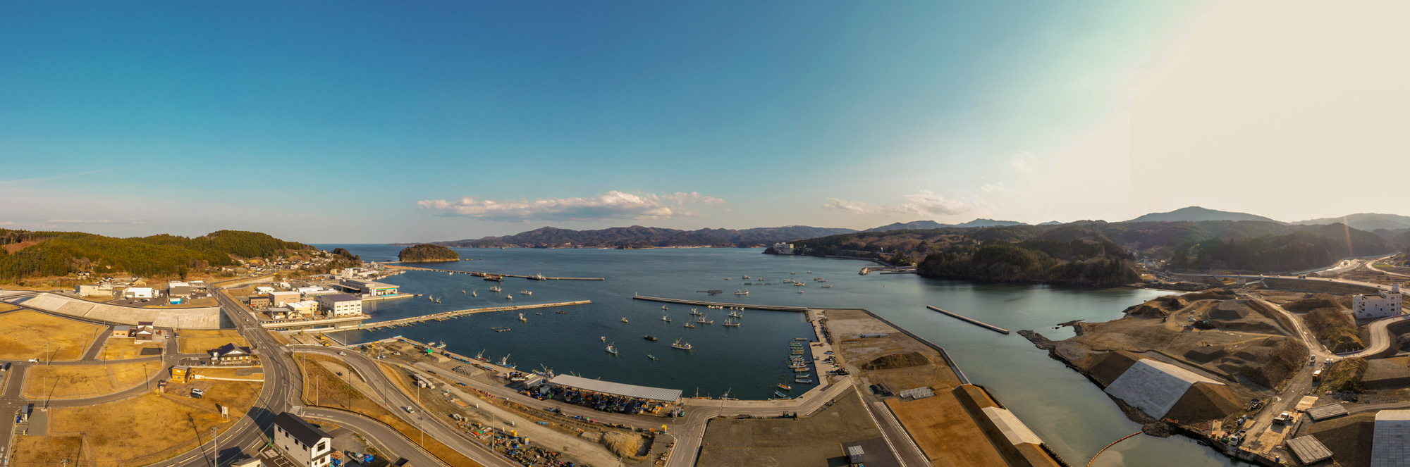 港に複数の船が停泊し、周囲には道路や建物、工事現場が広がり、背景には山々が連なり、青空の下で穏やかな雰囲気が漂う沿岸地域を上空から捉えた写真