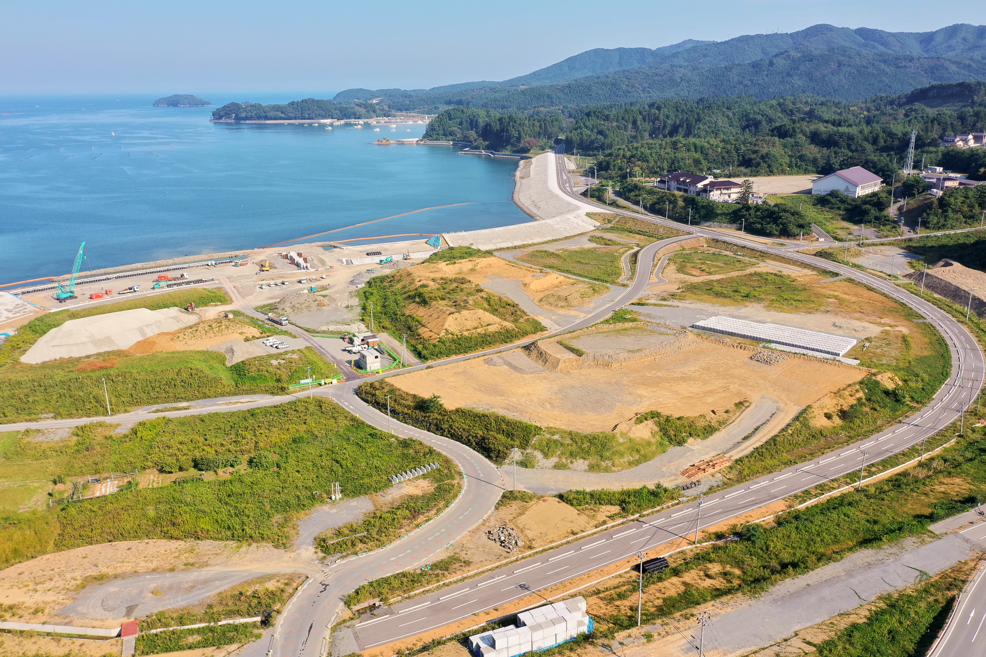 青い海と湾に囲まれた地域で、防潮堤と埋め立て工事が進む海岸沿いの風景を中心に、背後にある学校施設と山々を背景に収めた海岸再開発の様子の写真
