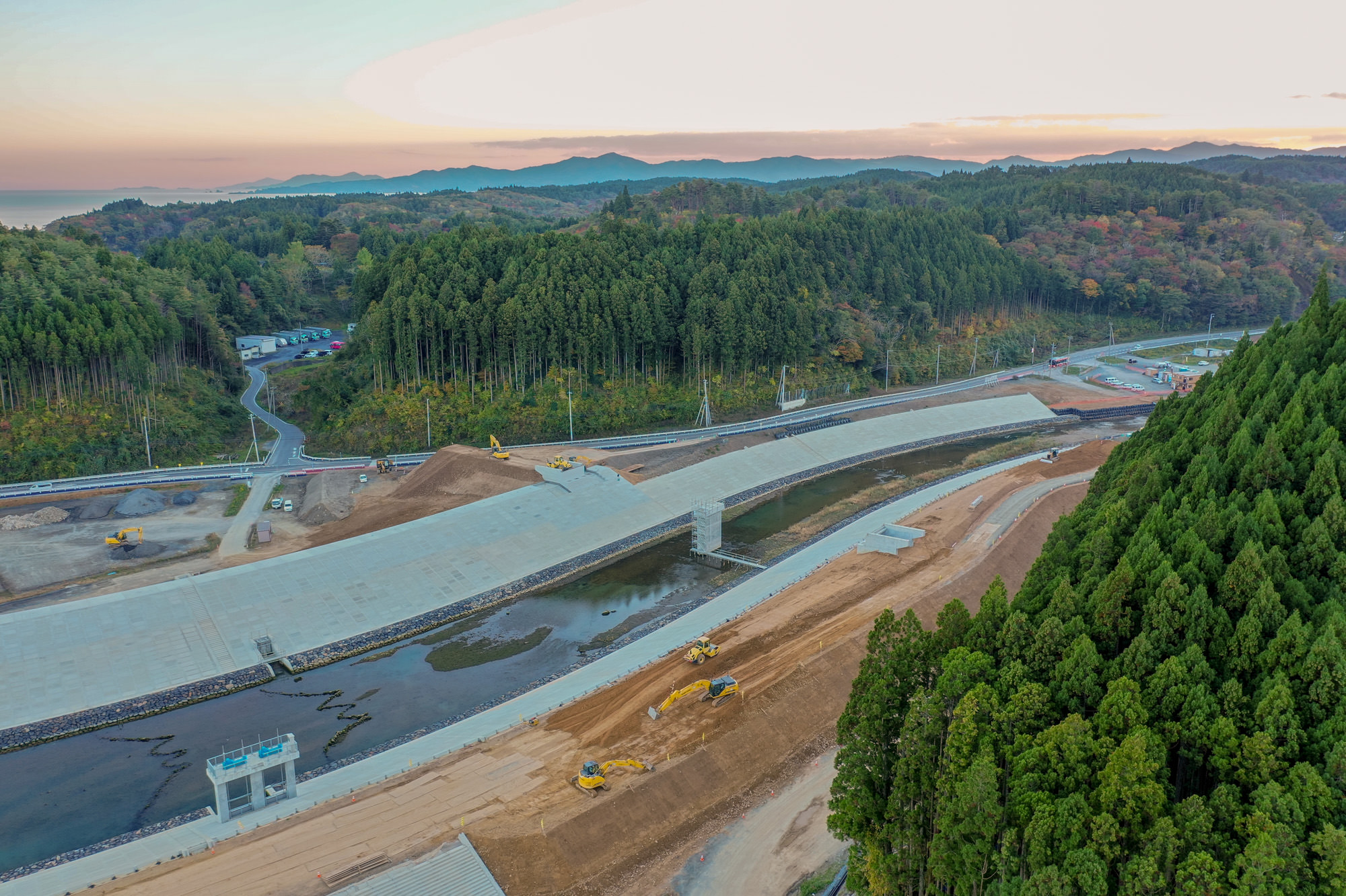 山間部に広がる大規模な建設現場で、川の上に橋が架けられており、その周囲には緑豊かな木々と複数の重機が配置されている様子が見える写真