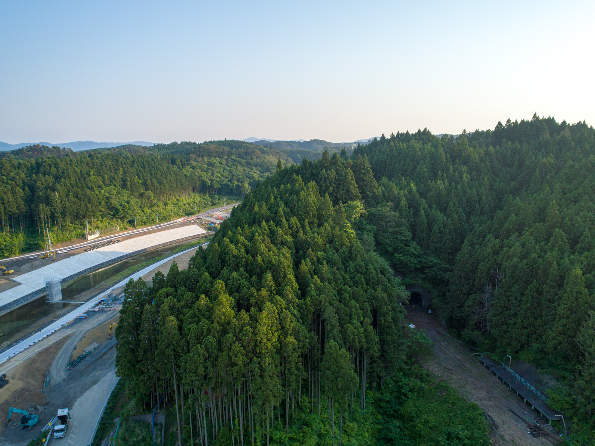 密集した森林が広がる地域を上空から撮影したもので、左側には風景に沿って曲がる建設中の道路があり、右側には木々に囲まれたトンネルの入口、背景には緑に覆われた丘と晴れた空が広がる風景の写真