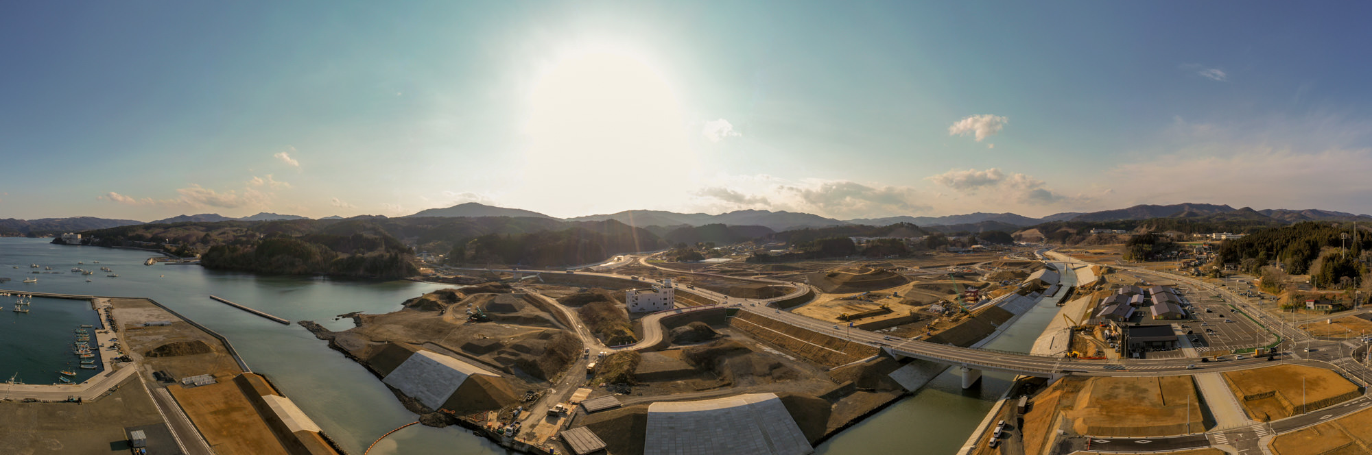 開発中または工事中の海沿いの地域を広く見渡すように撮影しており、構造物や道路、水路が点在し、背景には丘や山が広がり、空には太陽が明るく輝いている沿岸地域の写真