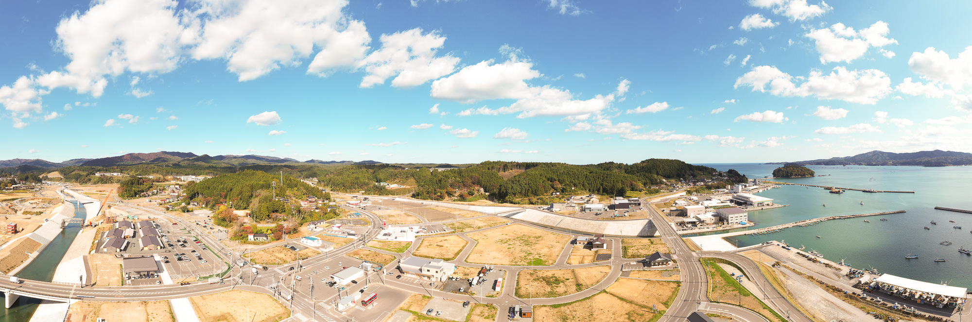 山側から海岸までを広く見渡せるパノラマ構図で、左側には整備された川と復興工事中の街並み、右側には漁港と防波堤、奥に広がる太平洋が見える復興中の港町全景の写真