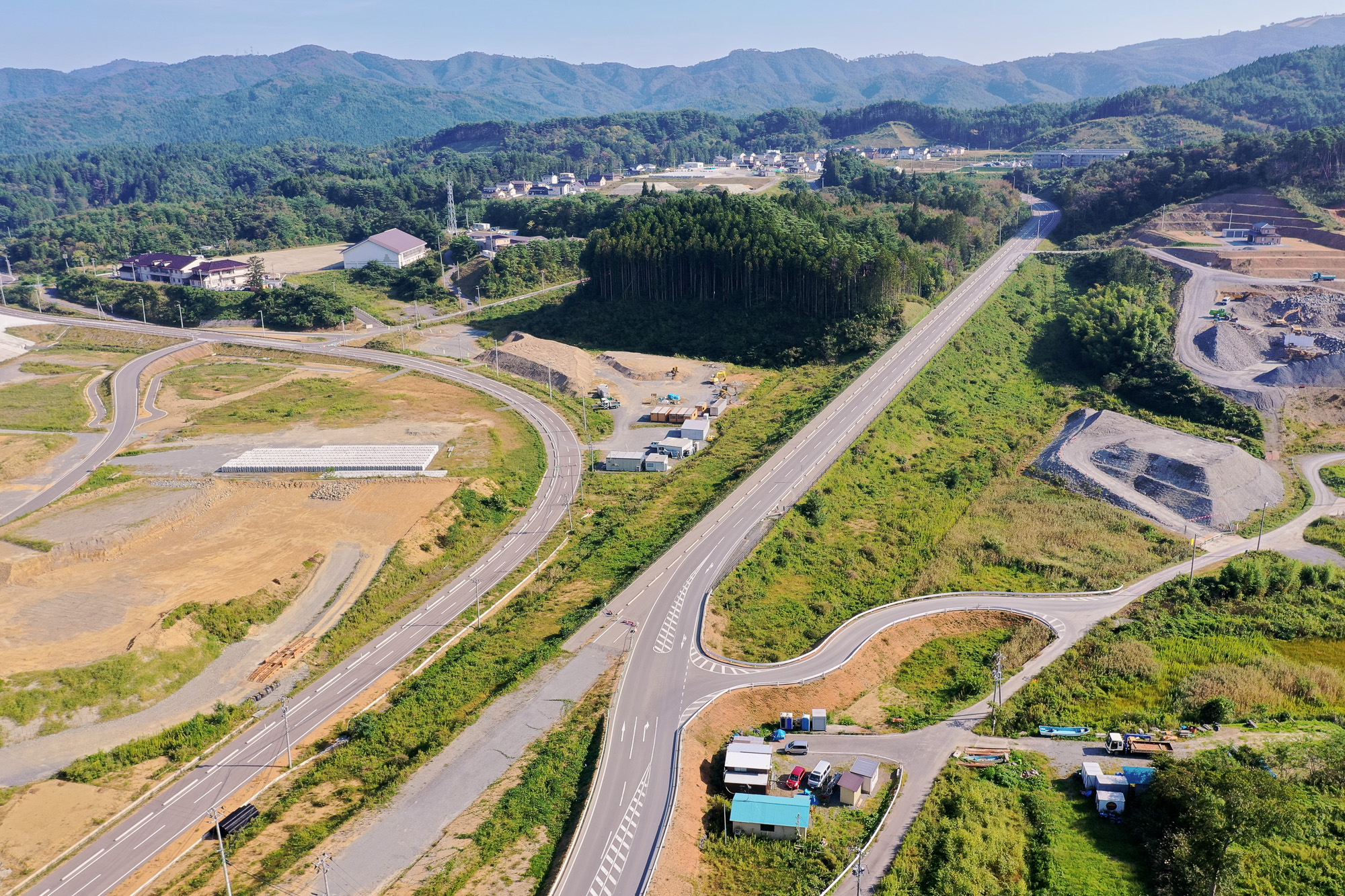 山あいを通る新しい道路が蛇行しながら延び、建設機材の置かれた敷地や住宅、学校らしき建物が見える、内陸部の再開発された道路周辺の様子の写真