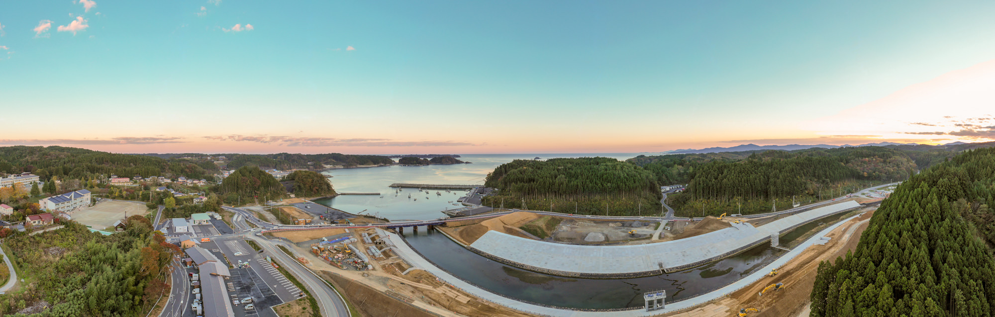 海に面した広大な風景で、背景には海が広がり、陸地が海に向かって突き出しており、手前には道路や橋、開発中と思われる施設があり、周囲には緑豊かな木々が広がり、空は青からオレンジに染まりつつあり日の出または日の入りの時間帯を示している写真