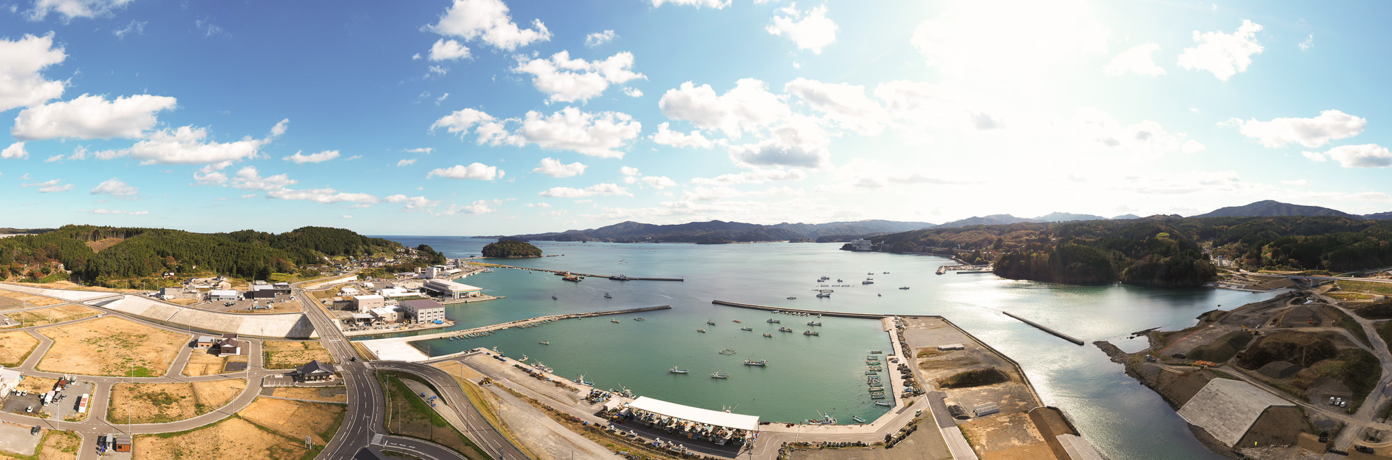 海に囲まれた湾の周囲に小型漁船が浮かび、左側には市街地と港、右側には整備中の護岸工事や道路が広がる風景を高所から広く見渡した写真