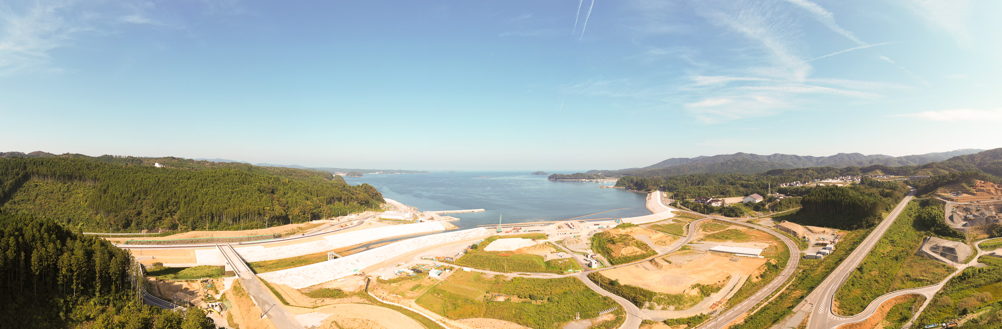 山と森に囲まれた海辺の町が見渡せる広角の空撮で、防潮堤の工事が進む海岸線と、内陸に延びる新しい道路、港の様子が確認できる三陸沿岸の復興状況の写真
