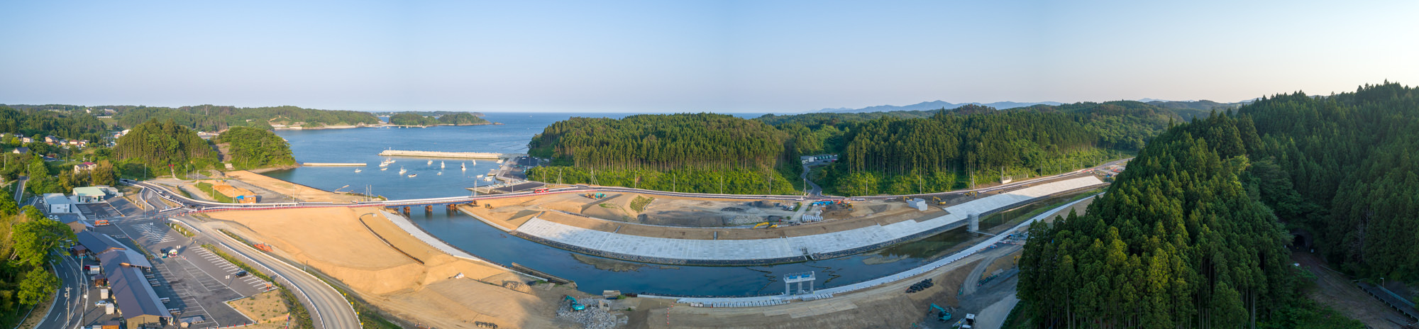 川が海へと流れ込む海岸地域を広く見渡したもので、川沿いでは治水や護岸工事が進められており、周囲には森林と丘が広がり、左側には道路や建物が見える人の暮らしと開発の様子がうかがえる風景のパノラマ写真