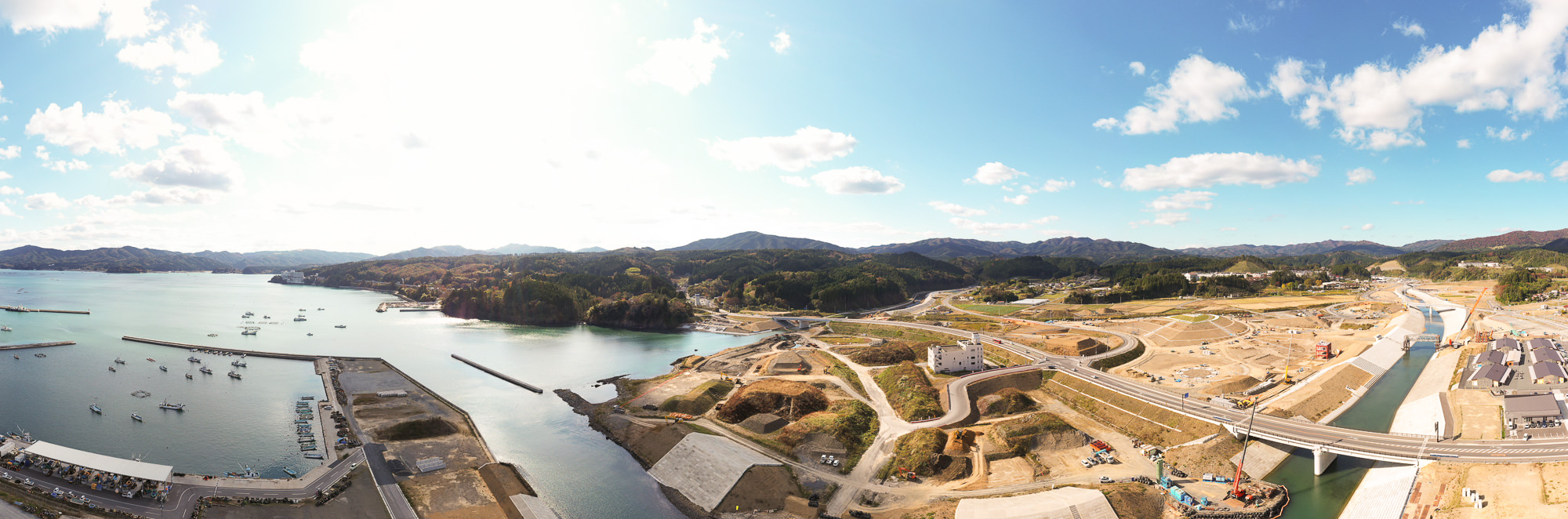 港から続く川の河口に沿って整備中の護岸と工事現場が広がり、奥には緑の山々と曲がりくねる道路が続く湾奥部のパノラマ写真