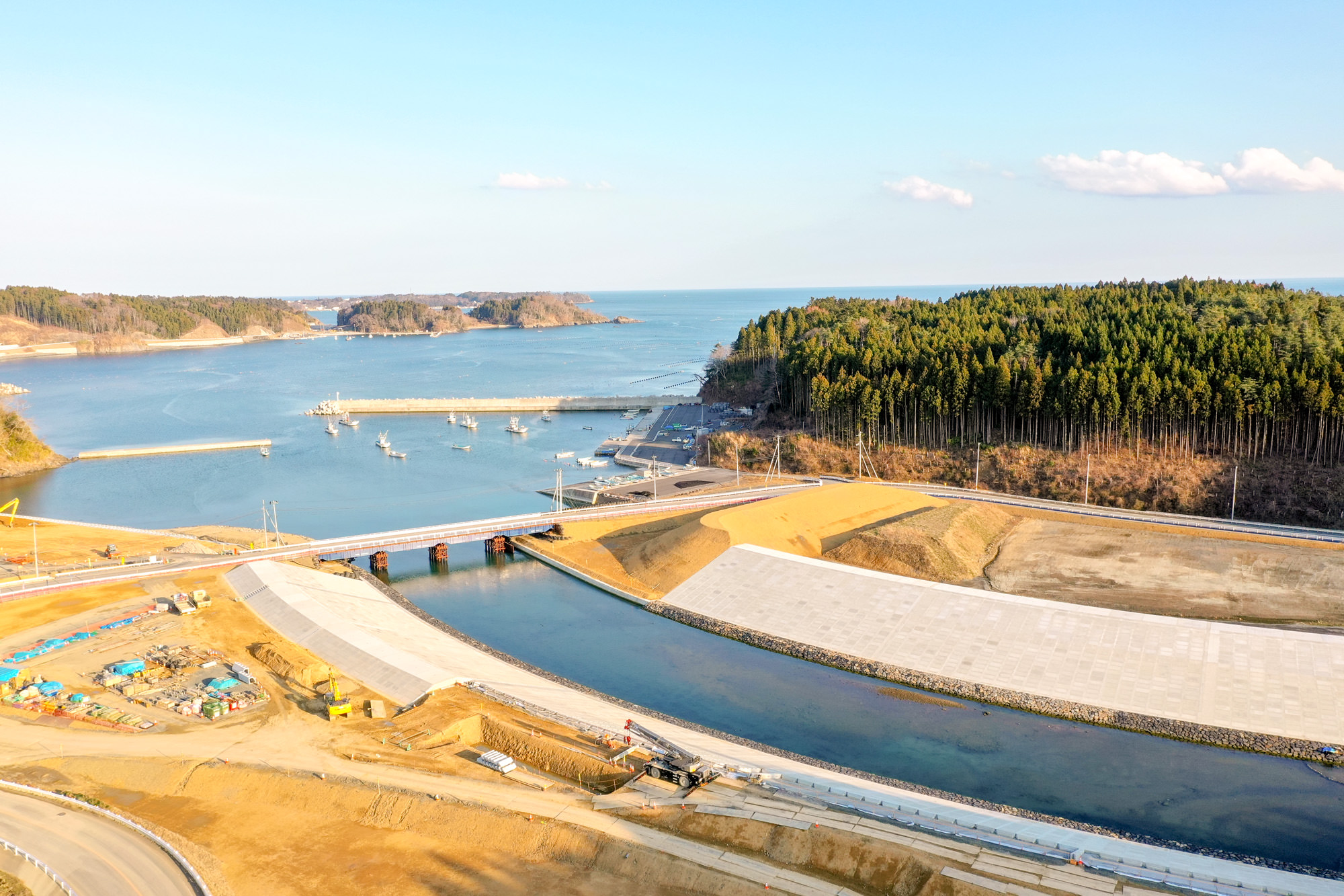 手前に川が流れ、その上に橋が架かり、川の両岸には工事中の様子が見られ、奥には森林と海が広がり、港湾には複数の船が停泊している海に面した港湾地域の写真
