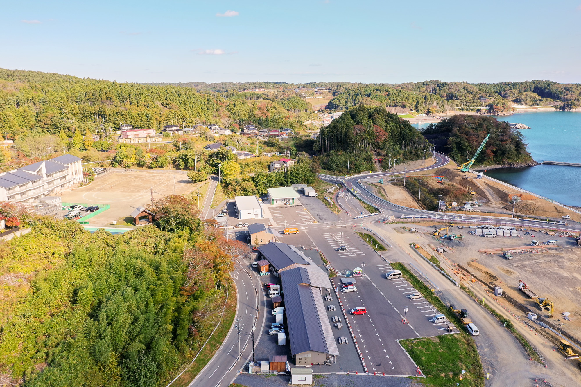 左下にある町の施設を上空から見下ろし、学校の校庭や駐車場、整備された建物が確認でき、背景には山の中に点在する民家が見える写真