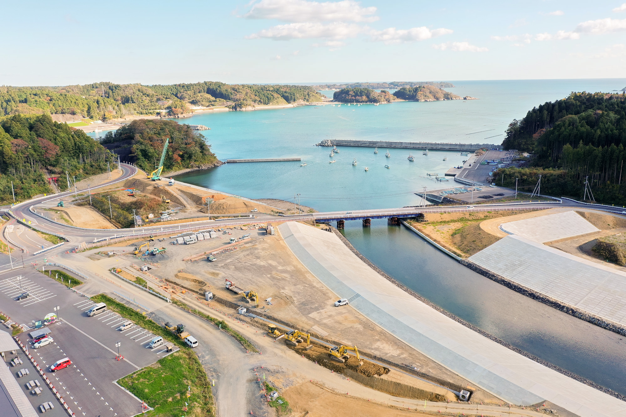 左側の漁港と右側の山に挟まれた場所に川が流れ、橋がかかり、周囲では重機による工事が進む様子を捉えた写真