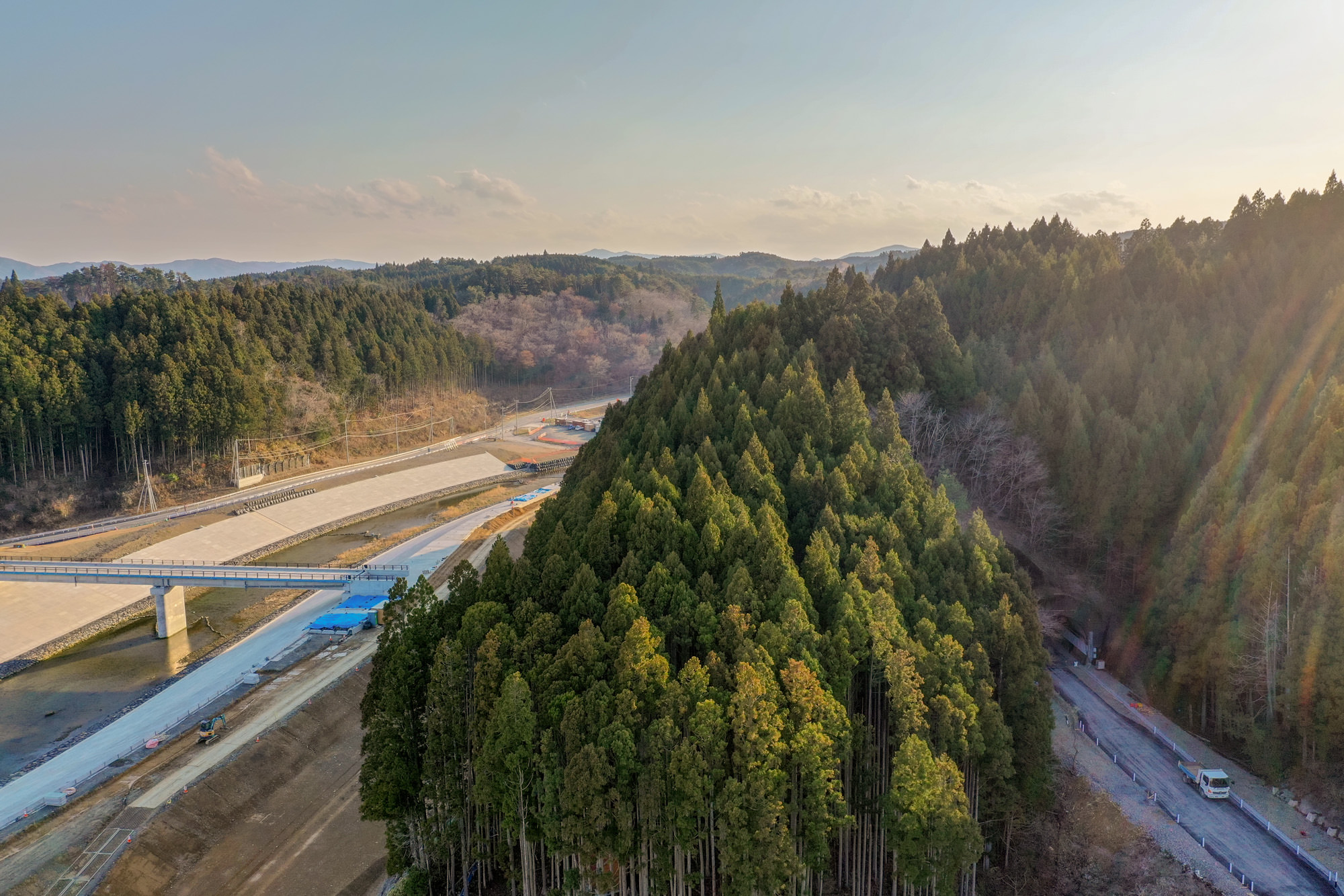 山間部の谷に沿って橋と道路が建設されており、その周囲には密集した森林が広がる様子を空から撮影した山間地域の写真