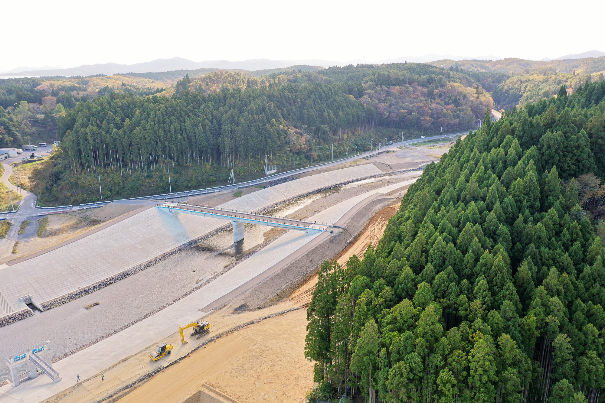 整備された川と両岸の護岸壁、その先に続く道路と山林、奥に連なる山並みが見える写真