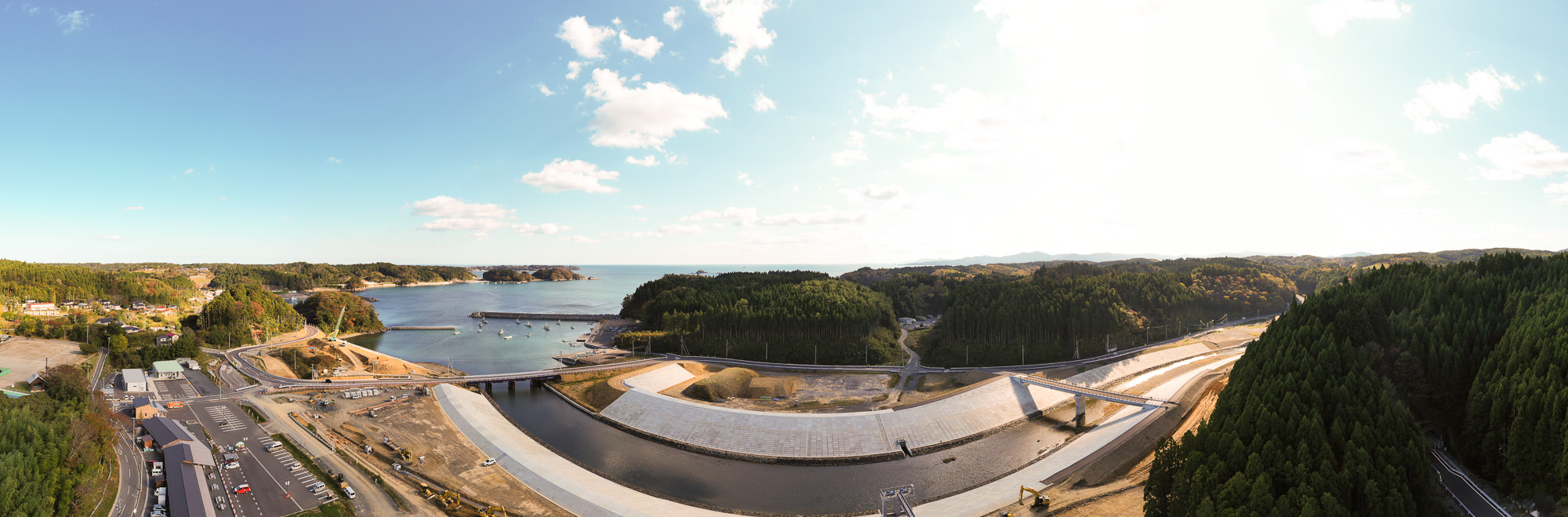 小さな漁港と周囲を囲む防波堤、その背後に広がる松林の丘と山々、中央には川と大規模な護岸工事現場、左手前には町の建物が並び、青空が広がる風景の写真