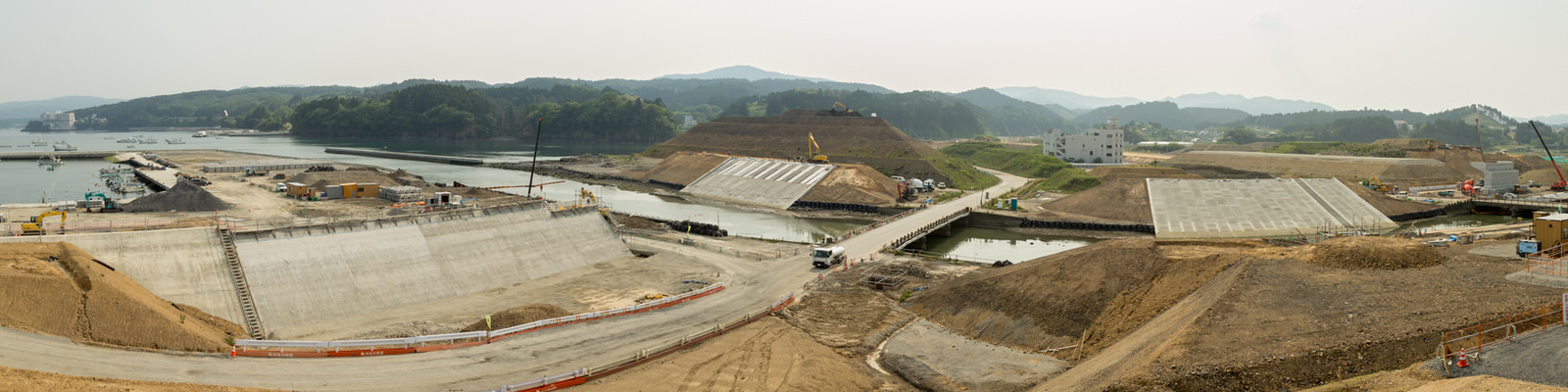 港へ流れる川沿いに面した広い工事現場で、複数のコンクリート構造物が建設中であり、橋が水の上をまたいで現場をつなぎ、クレーンやトラックなどの重機が稼働していて、背景には霞がかった空の下に森林に覆われた丘や山が広がっているパノラマ写真