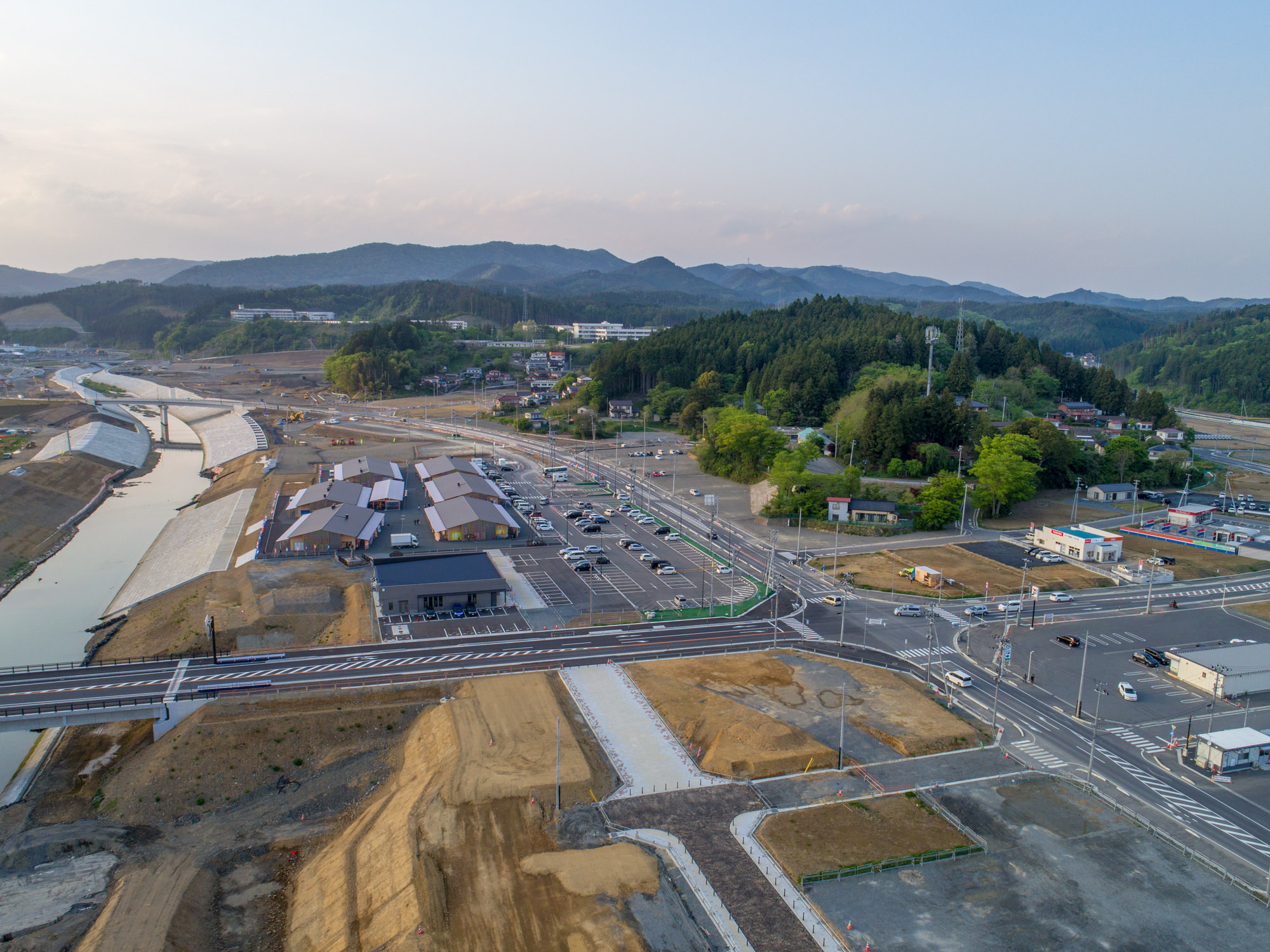 手前に複数の建物や駐車場、道路が広がり、左側には堤防に囲まれた川が流れ、背景には森林に覆われた丘や山々が広がる、晴れた空の下で撮影された農村地域の上空からの風景の写真