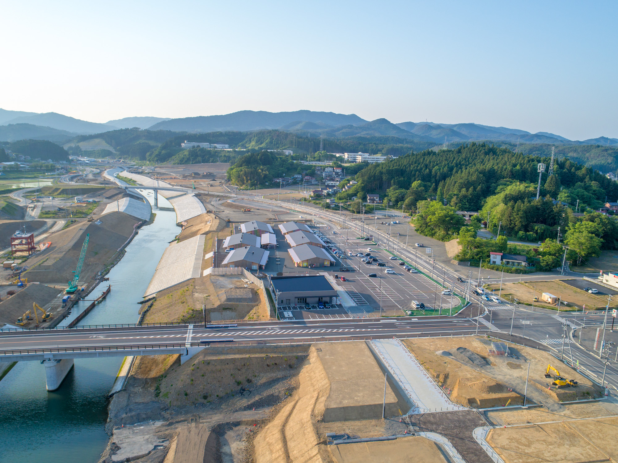 川の近くにある建設現場を上空から撮影したもので、手前に橋が架かり、中央には建物や駐車場があり、開発中の様子が見られ、背景には森林に覆われた丘と山々、雲のない青空が広がる風景の写真
