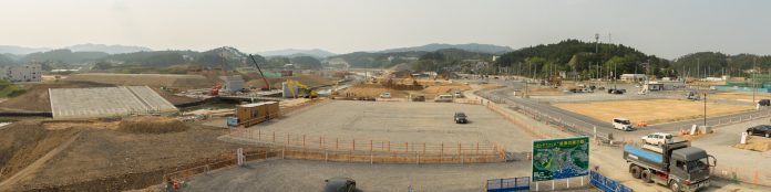 広大な工事現場の全景で、複数の重機が土を掘り起こしたり整地作業を行っており、地面は茶色く掘り返され、周囲には緑豊かな山々が連なっていて、自然と人工の風景が対比的に広がっている様子のパノラマ写真