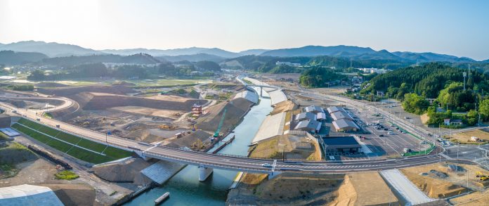 広範囲にわたる建設現場を上空から撮影したもので、中央に川と橋があり、右側に駐車場と建物、左側に道路と緑地、背景には山々が広がる自然と人工構造物が調和した風景の写真