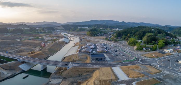 川に架かる大きな橋と、その周辺で進められている道路や施設の建設現場を上空から捉え、遠くには緑豊かな山々が広がっている様子を写した風景のパノラマ写真